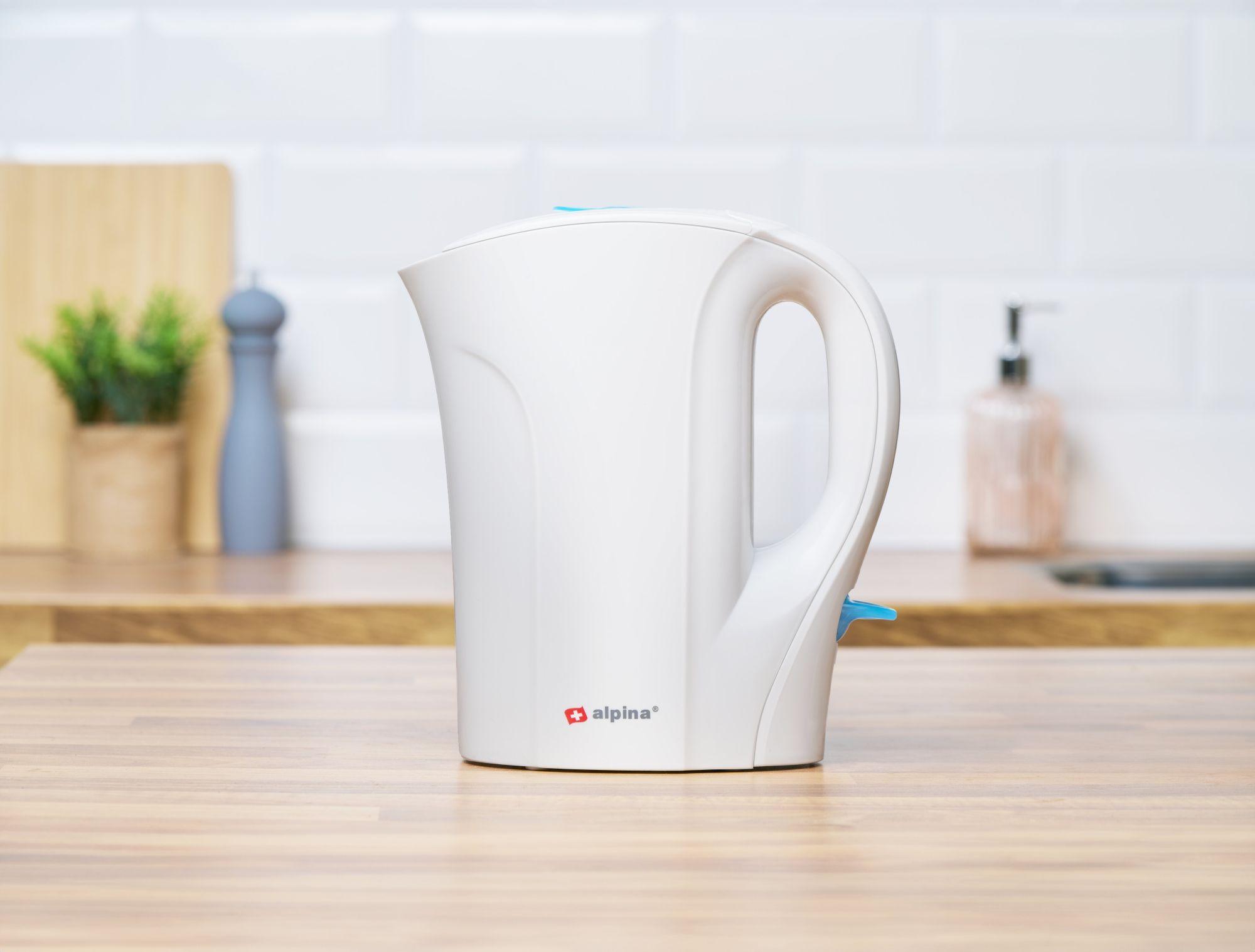 Biały czajnik Alpina stojący bokiem na drewnianym blacie. W tle widać kuchnie.