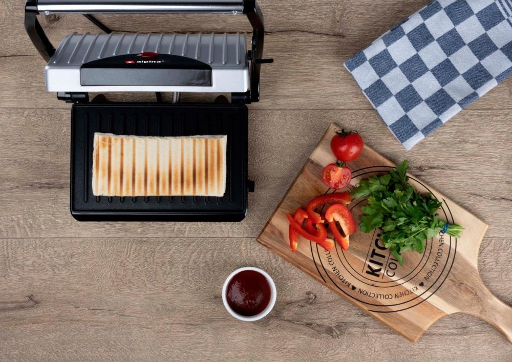 Otwarty opiekacz z kanapka w środku. Obok jest deska z pokrojonymi warzywami.