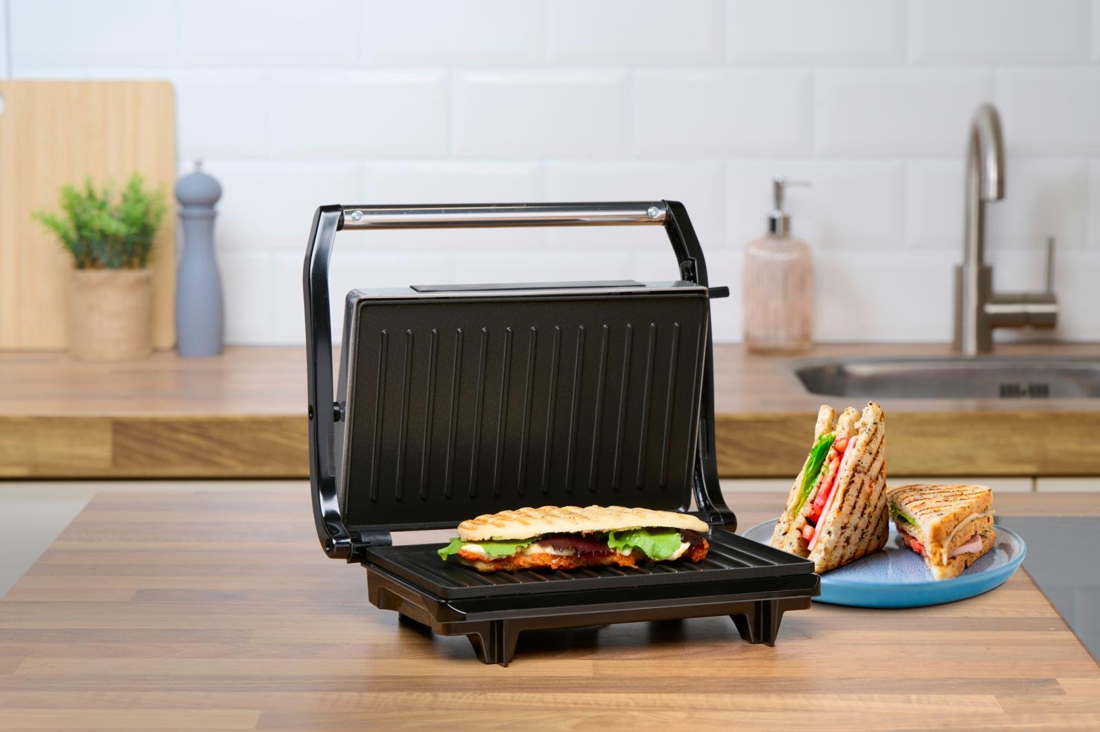 Otwarty grill w środku leży kanapka. Obok urządzenia stoi talerz z kanapkami.