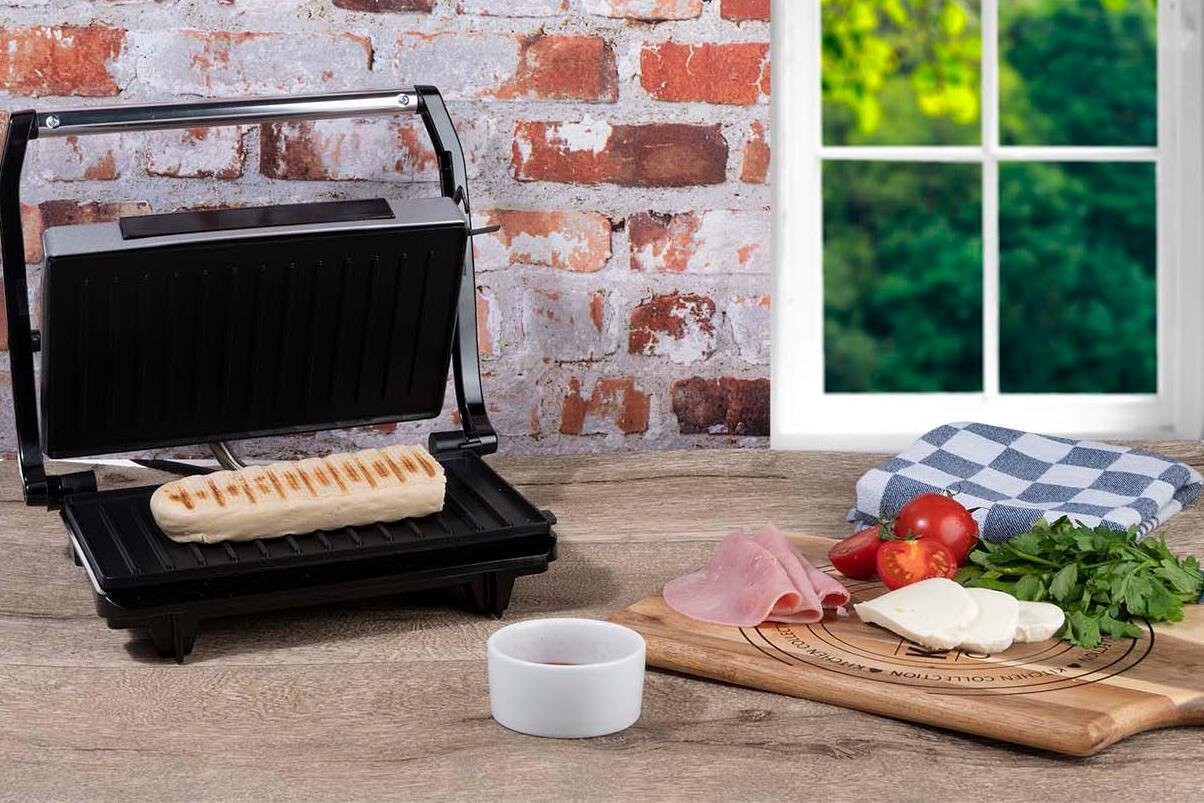 Otwarty opiekacz z kanapka w środku. Obok jest deska z pokrojonymi warzywami. Urządzanie stoi w kuchni na blacie.