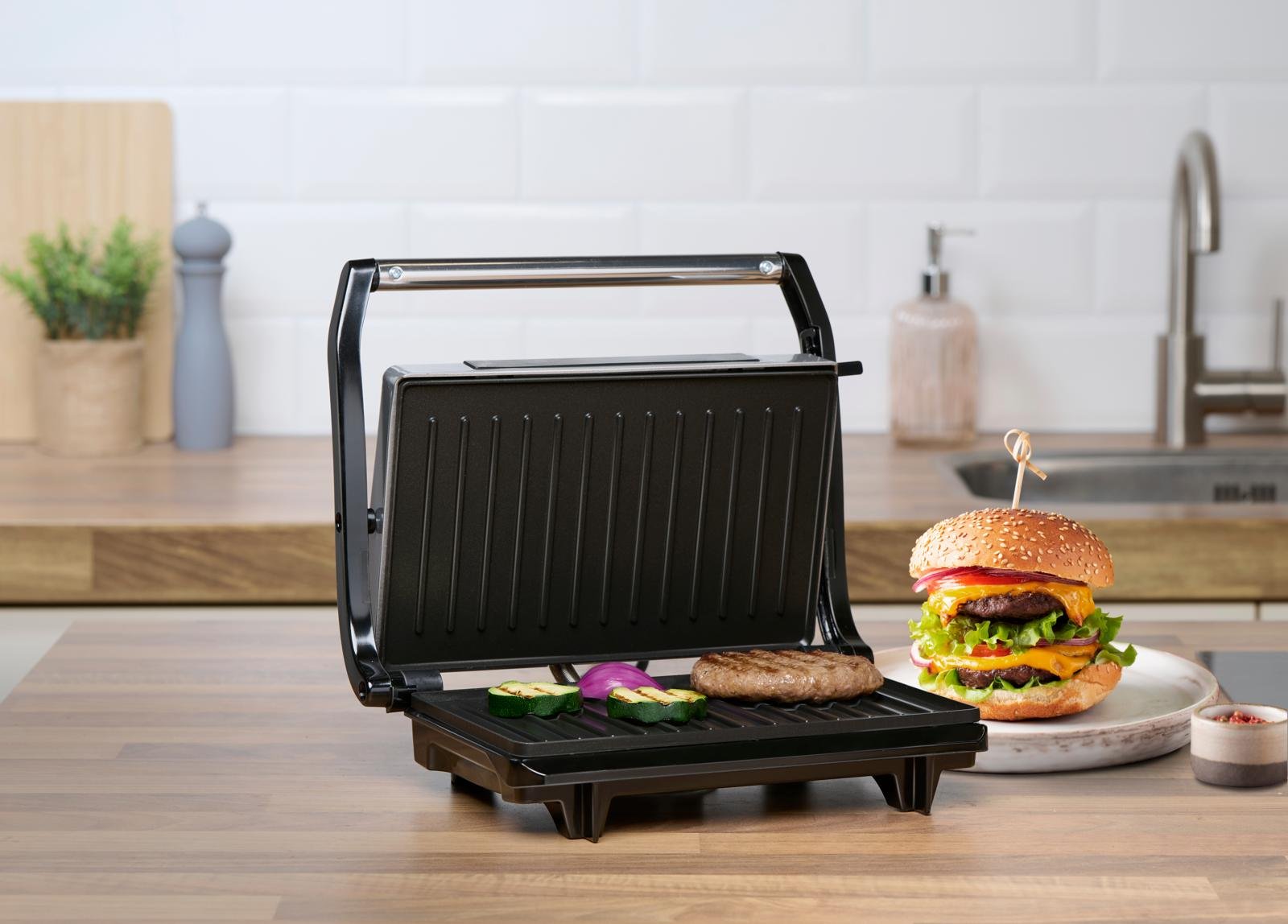 Otwarty opiekacz z kanapka w środku. Obok jest talerz z burgerem. Urządzanie stoi w kuchni na blacie.