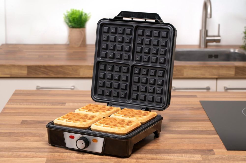 Otwarta czarna gofrownica Alpina stojąc na blacie kuchennym. W środku smażą się 4 gofry.